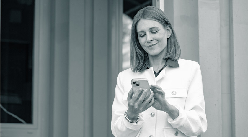 Eine Frau mit schulterslangen, hellen Haaren und einem schicken Mantel blickt zufrieden auf ihr Smartphone.